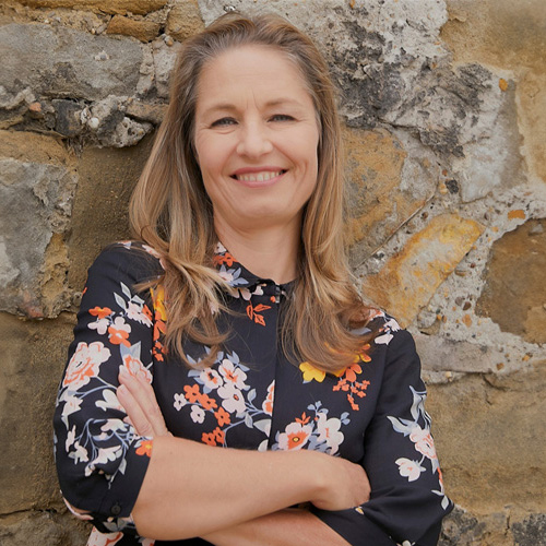 Portraitfoto von Alexandra Fiedler-Lehmann. Die Inhaberin der "Guten Agentur" hat lange blonde Haare und trägt eine geblümte Bluse. Sie steht mit verschränkten Armen vor einer Steinmauer und lächelt Richtung Kamera.